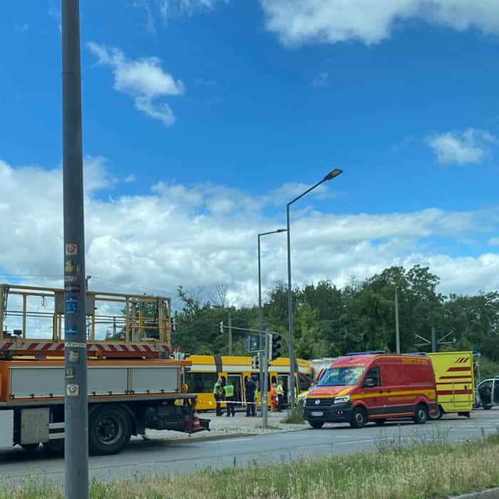Blick aus einem Fahrzeug auf eine Unfallstelle in Dresden: Ein orangefarbenes Rettungsfahrzeug, ein roter Einsatzleitwagen und die Polizei stehen an einer gelben Straßenbahn. Rechts unten spiegelt sich die Straße im Außenspiegel.