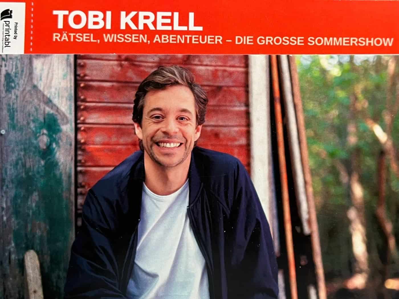 Ein rotes Ticket für die Sommershow von Tobi Krell liegt auf einem Holztisch. Auf der Eintrittskarte ist ein Foto des lächelnden Moderators vor einer Holzhütte abgebildet. Das aufgedruckte Veranstaltungsdatum zeigt den 27.06.2026 in Dresden.