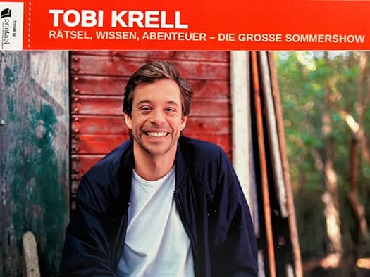 Ein rotes Ticket für die Sommershow von Tobi Krell liegt auf einem Holztisch. Auf der Eintrittskarte ist ein Foto des lächelnden Moderators vor einer Holzhütte abgebildet. Das aufgedruckte Veranstaltungsdatum zeigt den 27.06.2026 in Dresden.