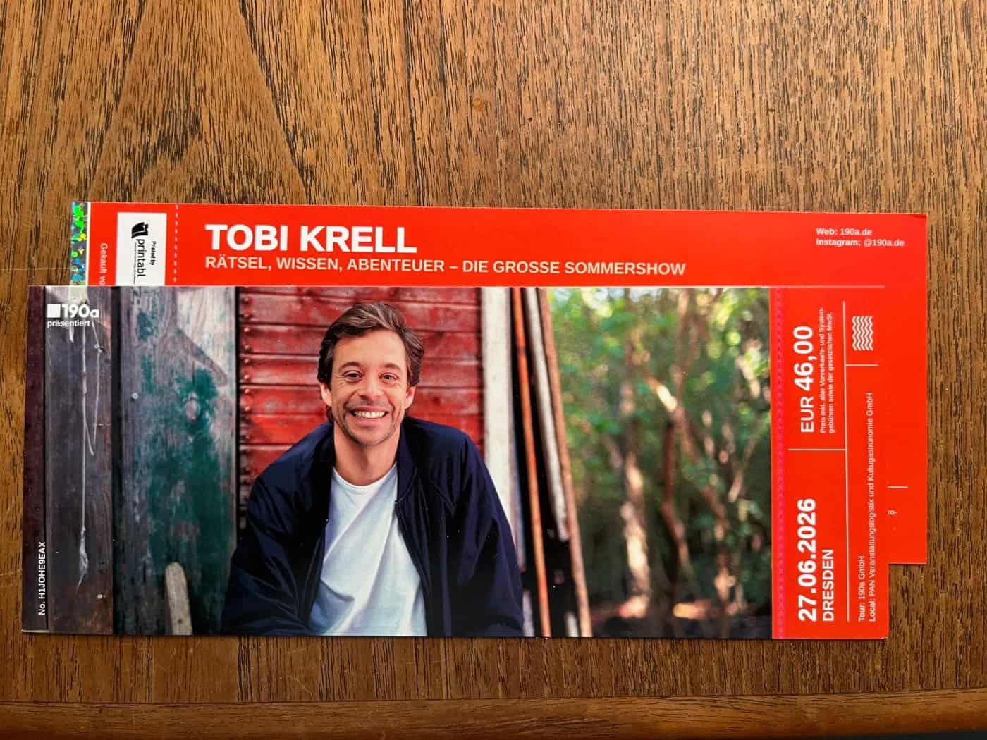 Ein rotes Ticket für die Sommershow von Tobi Krell liegt auf einem Holztisch. Auf der Eintrittskarte ist ein Foto des lächelnden Moderators vor einer Holzhütte abgebildet. Das aufgedruckte Veranstaltungsdatum zeigt den 27.06.2026 in Dresden.