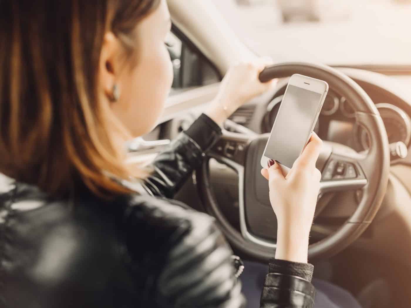 Frau hält ein Smartphone in der Hand, während sie am Steuer eines Autos sitzt und auf das Display schaut.