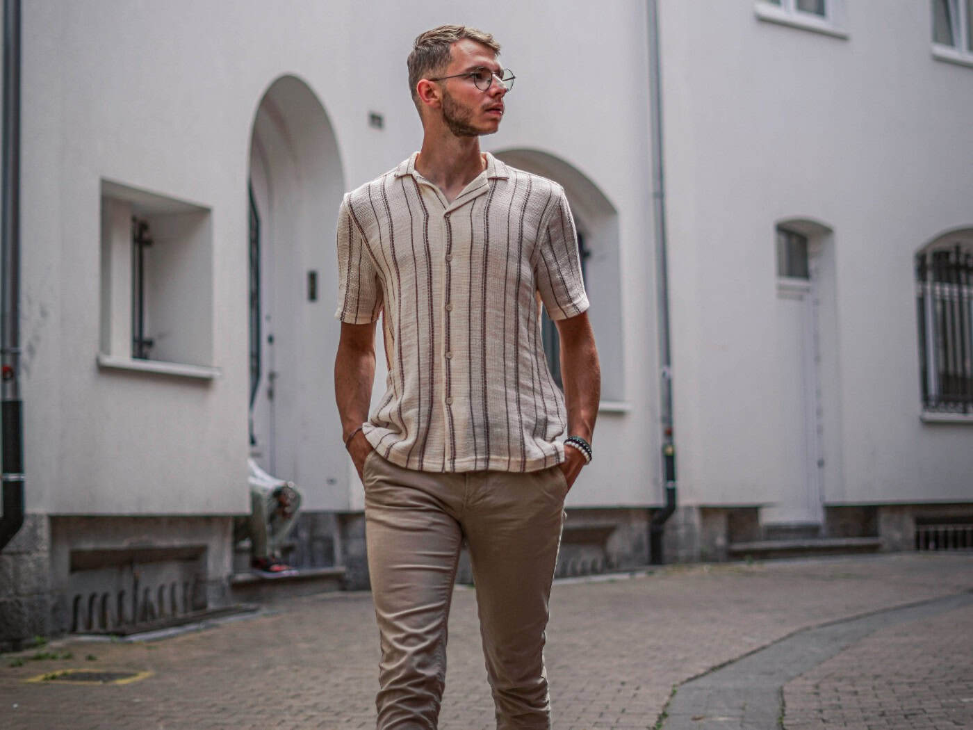 Mann in beigefarbenen Leinenhosen und gestreiftem Hemd geht entspannt durch eine Gasse – perfektes Sommeroutfit für Hitze und Alltag.