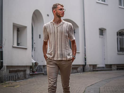 Mann in beigefarbenen Leinenhosen und gestreiftem Hemd geht entspannt durch eine Gasse – perfektes Sommeroutfit für Hitze und Alltag.