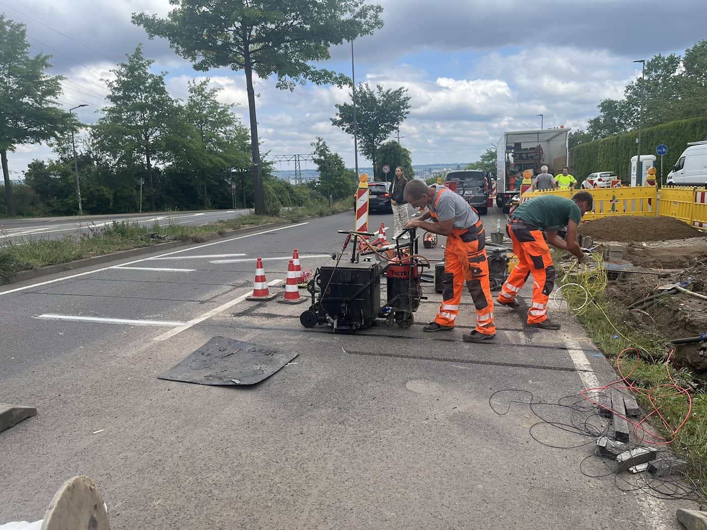 Zwei Bauarbeiter in orangefarbener Arbeitskleidung arbeiten an einer Baustelle auf einer gesperrten Straße. Ein Arbeiter steht neben einer Asphaltfräse, der andere hantiert mit gelben Kabeln an einer Grube am Straßenrand.