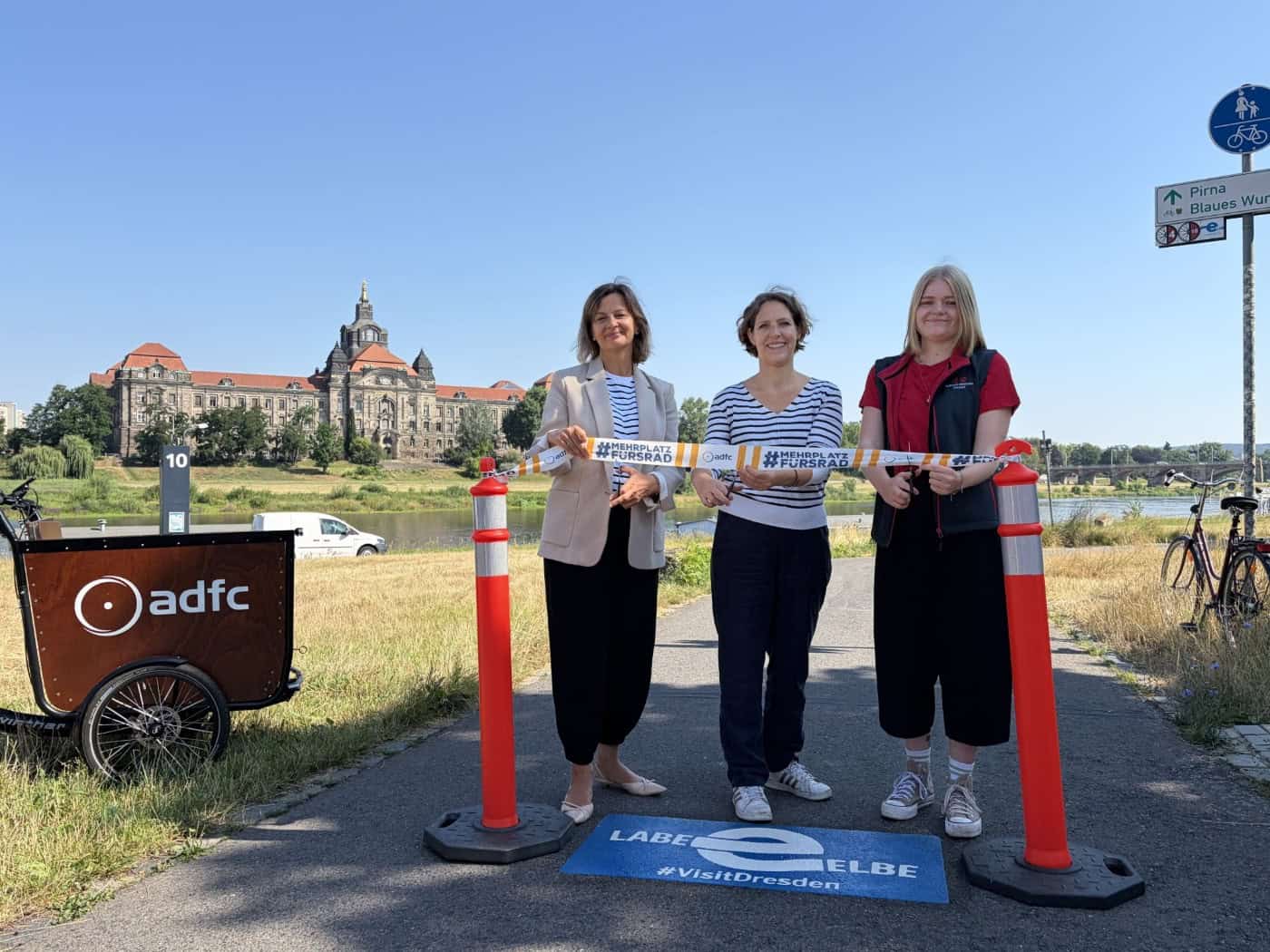 Karla Kallauch, Juliane Mostertz und Jule Thöming (v.l.n.r.) schneiden ein Band für das neue Elberadweg-Projekt in Dresden durch. Links steht ein ADFC-Lastenrad, im Hintergrund das Sächsische Finanzministerium bei blauem Himmel.
