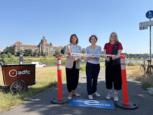 Karla Kallauch, Juliane Mostertz und Jule Thöming (v.l.n.r.) schneiden ein Band für das neue Elberadweg-Projekt in Dresden durch. Links steht ein ADFC-Lastenrad, im Hintergrund das Sächsische Finanzministerium bei blauem Himmel.