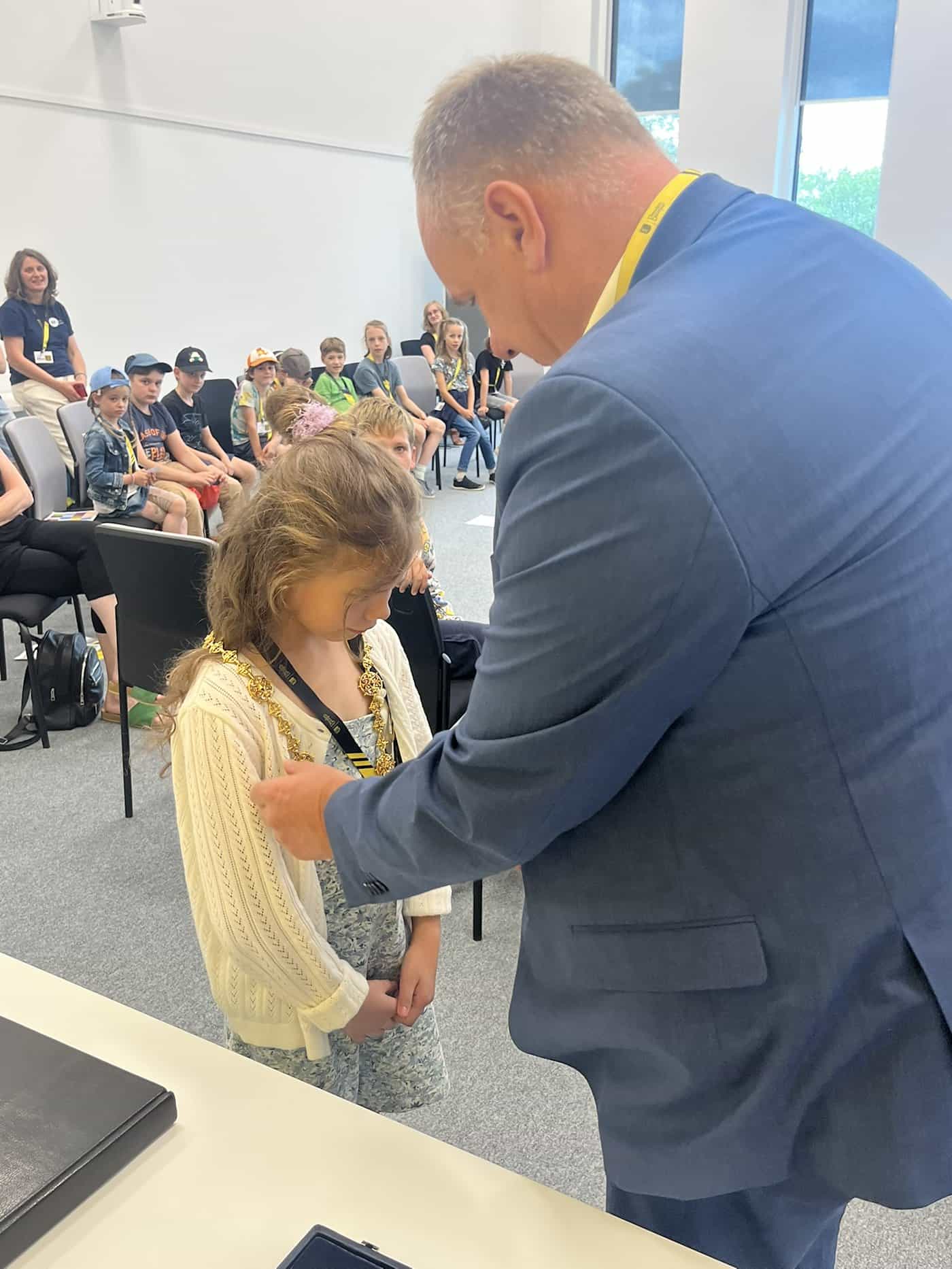 Oberbürgermeister Dirk Hilbert legt von der Seite einer schüchternen Grundschülerin in weißer Strickjacke die schwere goldene Amtskette Dresdens um den Hals. Im Hintergrund sitzen weitere Kinder.