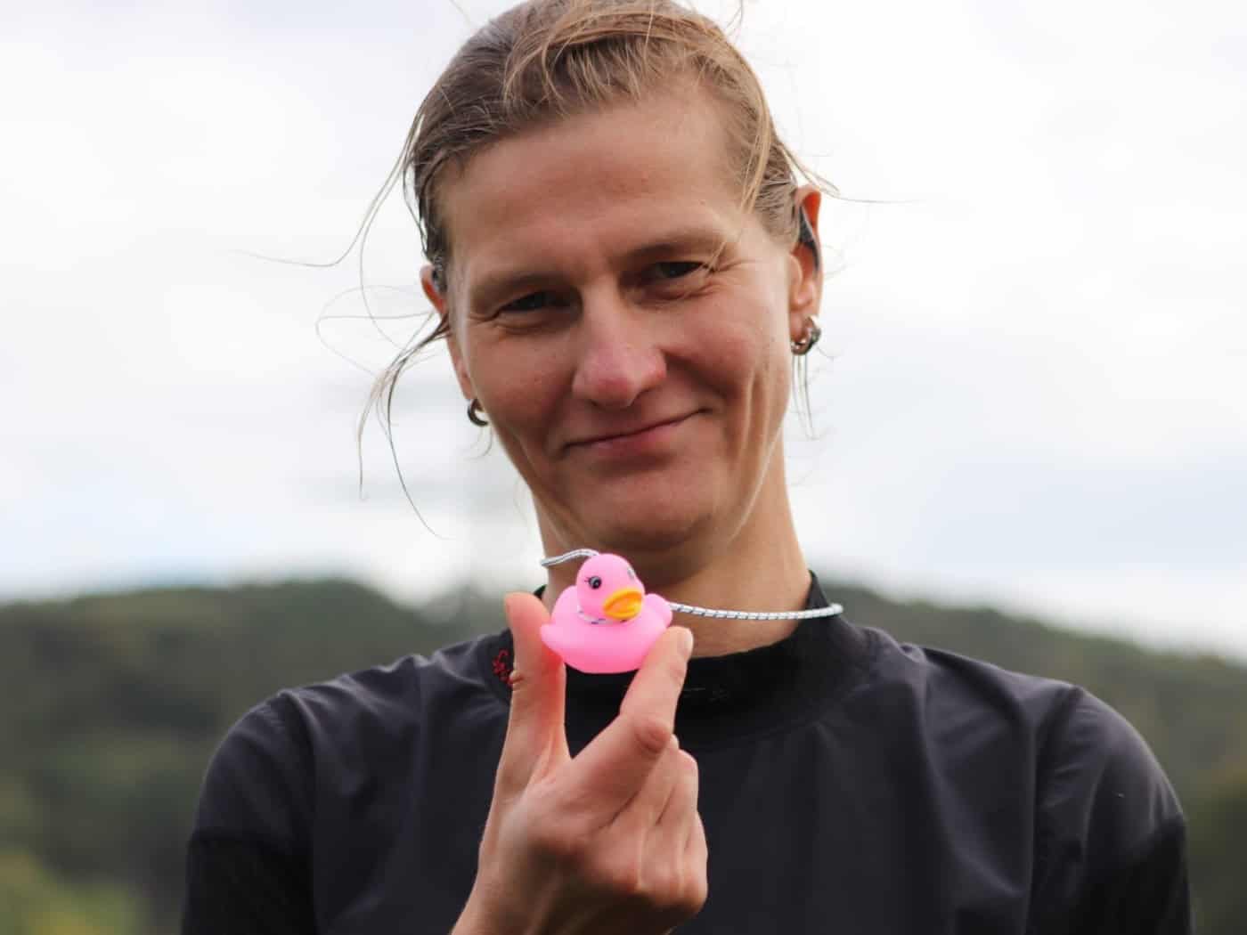 Nahaufnahme einer lächelnden Frau mit hochgebundenen Haaren im dunklen Sportshirt, die eine kleine, pinke Quietscheente an einer elastischen Schnur vor sich in der Hand hält. Der Hintergrund ist hell und unscharf.