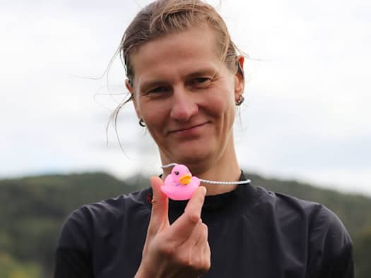 Nahaufnahme einer lächelnden Frau mit hochgebundenen Haaren im dunklen Sportshirt, die eine kleine, pinke Quietscheente an einer elastischen Schnur vor sich in der Hand hält. Der Hintergrund ist hell und unscharf.