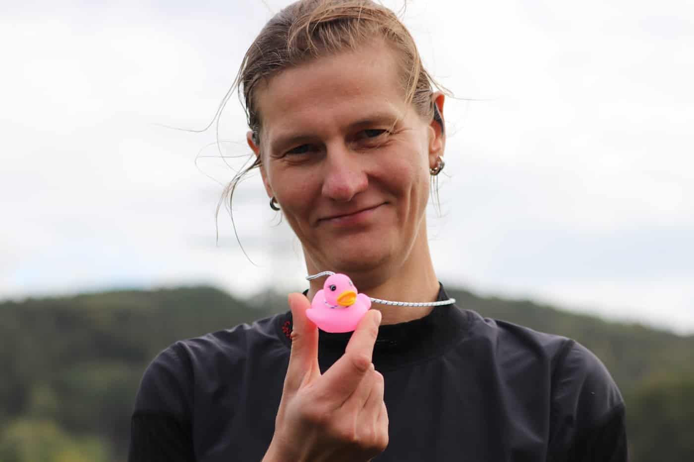 Nahaufnahme einer lächelnden Frau mit hochgebundenen Haaren im dunklen Sportshirt, die eine kleine, pinke Quietscheente an einer elastischen Schnur vor sich in der Hand hält. Der Hintergrund ist hell und unscharf.