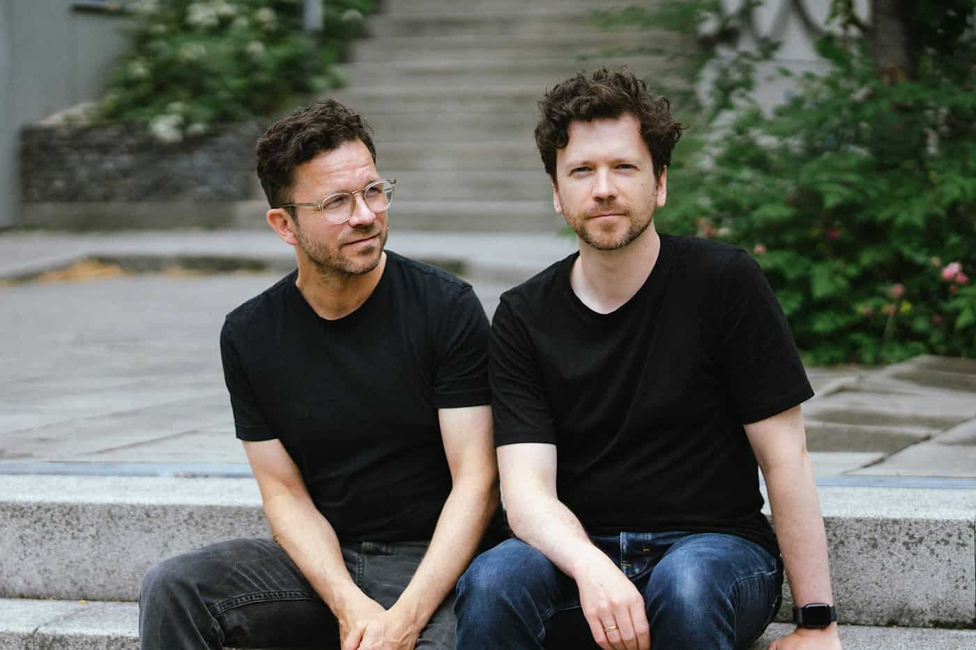 Die Architekten Patrick Hesse und Vincent Rasser von Neoform Architektur Porträt der beiden Architekten Patrick Hesse und Vincent Rasser. Sie tragen schwarze T-Shirts und sitzen nebeneinander auf den grauen Steinstufen einer Außentreppe. Einer blickt nach links zur Seite, der andere direkt in die Kamera.