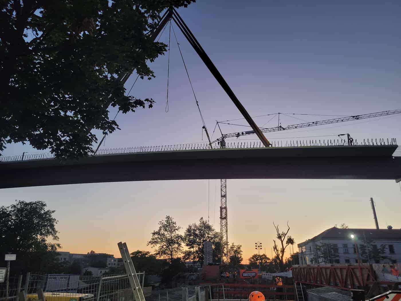 Ein Brückenträger schwebt am Kran über der Baustelle Fabricestraße Dresden im Abendlicht Blick von unten gegen den dämmernden Abendhimmel: Ein langer, dunkler Brückenträger hängt waagerecht an Tragseilen eines Krans. Links rahmen dunkle Blätter das Bild, im Hintergrund steht ein filigraner Baukran.