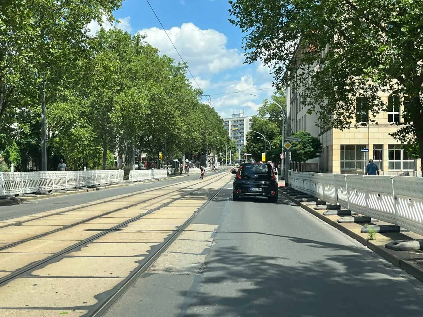 Blick auf die Güntzstraße in Dresden mit bereits aufgestellten weißen Baustellenzäunen entlang der Straßenbahngleise. Ein schwarzes Auto fährt auf der verbleibenden Spur, gesäumt von grünen Bäumen.