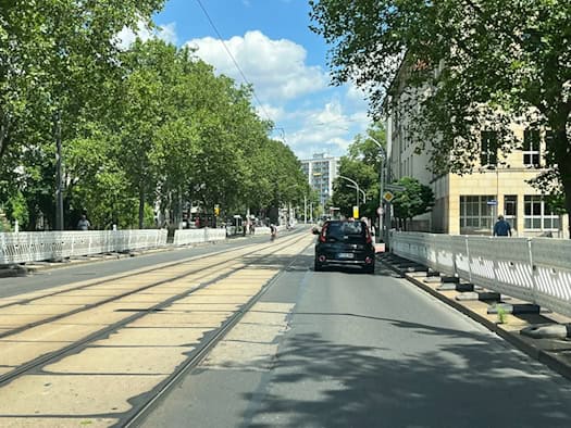 Blick auf die Güntzstraße in Dresden mit bereits aufgestellten weißen Baustellenzäunen entlang der Straßenbahngleise. Ein schwarzes Auto fährt auf der verbleibenden Spur, gesäumt von grünen Bäumen.