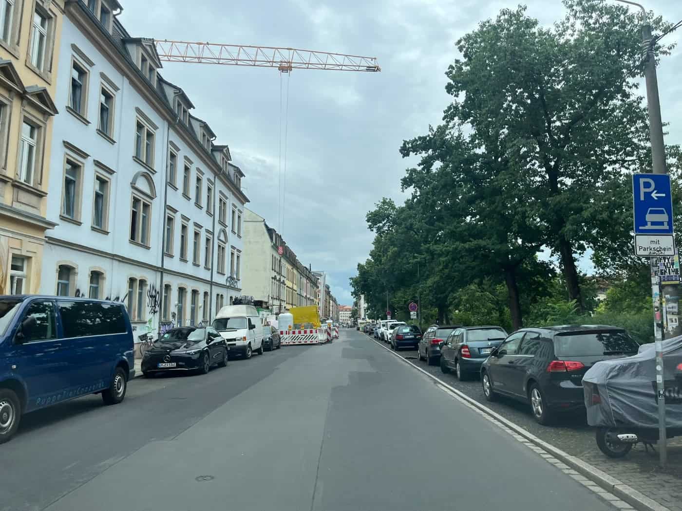 Eine Straße in Dresden mit geparkten Autos und Wohnhäusern links sowie Bäumen rechts. Am bewölkten Himmel ist ein großer gelber Baukran zu sehen, im Vordergrund rechts steht ein blaues Parkplatzschild.