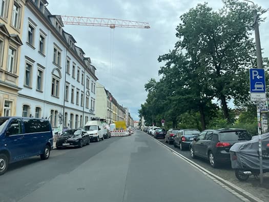Eine Straße in Dresden mit geparkten Autos und Wohnhäusern links sowie Bäumen rechts. Am bewölkten Himmel ist ein großer gelber Baukran zu sehen, im Vordergrund rechts steht ein blaues Parkplatzschild.