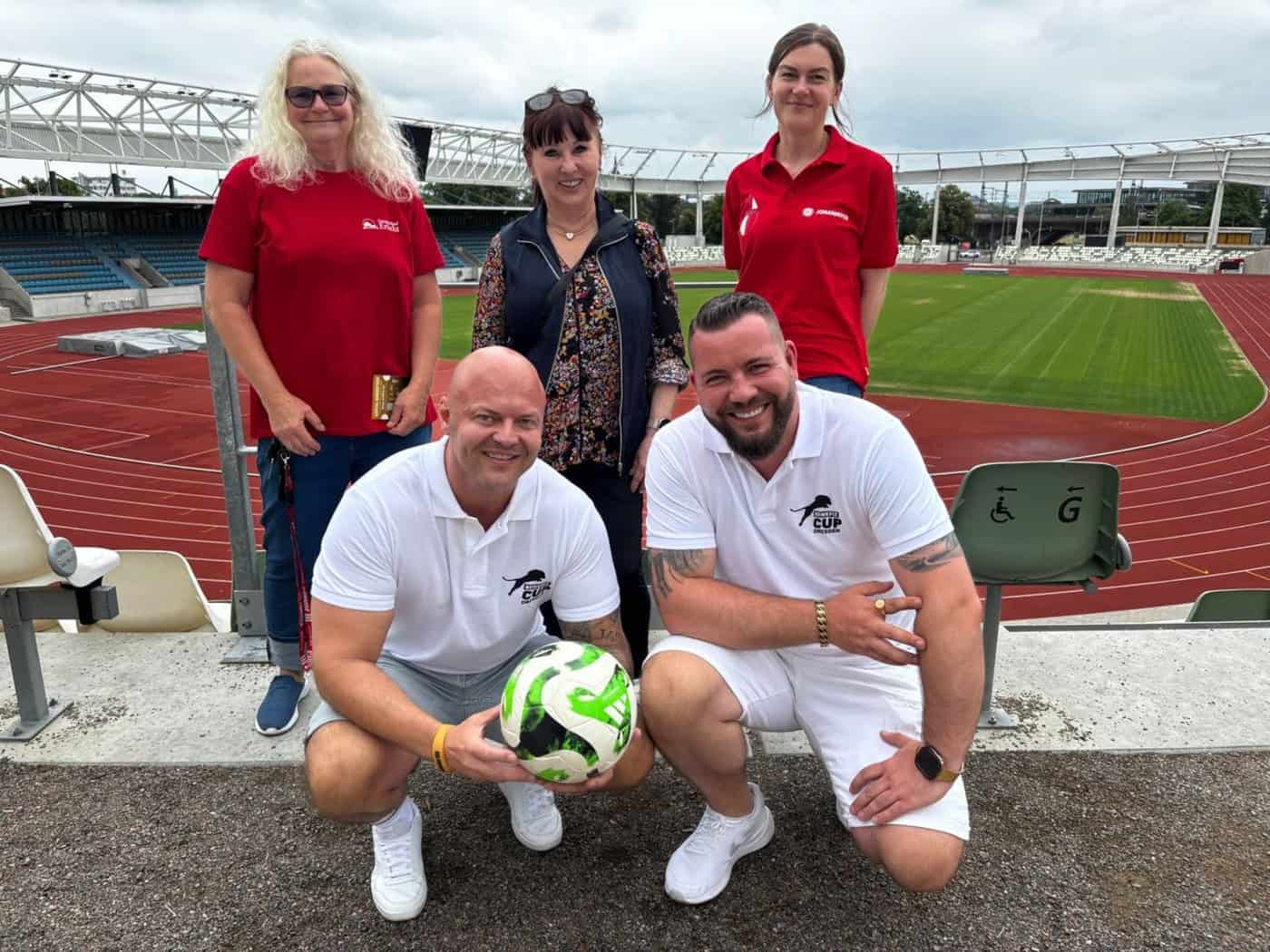 Fünf Personen posieren für ein Gruppenfoto auf der Tribüne des Heinz-Steyer-Stadions in Dresden. Zwei Männer knien im Vordergrund mit einem Fußball, dahinter stehen drei Frauen. Im Hintergrund ist das grüne Spielfeld zu sehen.