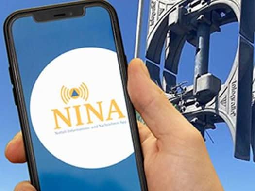 Eine Hand hält ein Smartphone im Vordergrund, auf dessen Display das kreisförmige Logo der NINA Warn-App zu sehen ist. Im unscharfen Hintergrund ist eine graue elektronische Sirene an einem Mast vor einem klaren, strahlend blauen Himmel montiert.