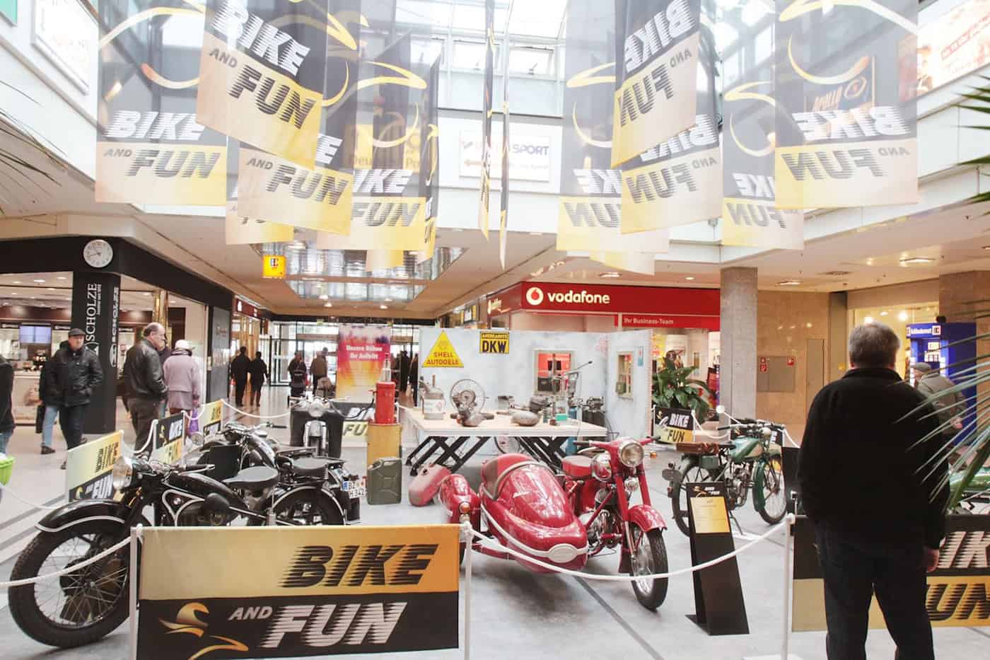 Ausstellungsfläche mit mehreren Motorrädern in einem Shopping-Center, darüber und als Absperrung Banner mit der Aufschrift "Bike and Fun"