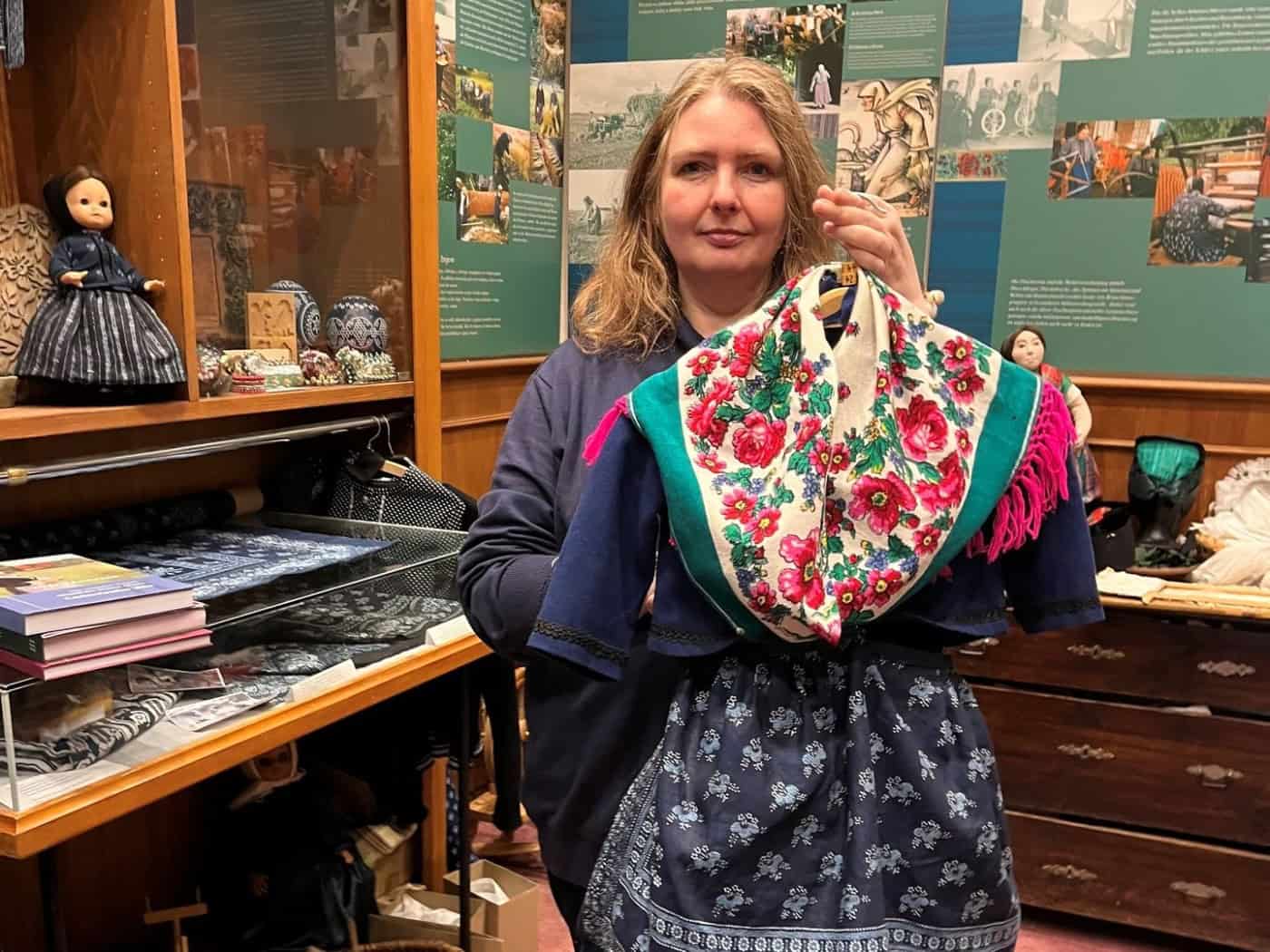 Eine Frau schaut in die Kamera und hält auf einem Bügel vor sich eine sorbische Tracht mit Blumenmotiven und Blaudruck.