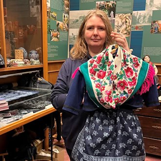 Eine Frau schaut in die Kamera und hält auf einem Bügel vor sich eine sorbische Tracht mit Blumenmotiven und Blaudruck.