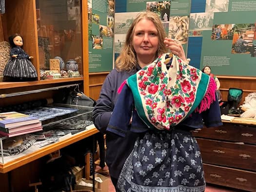 Eine Frau schaut in die Kamera und hält auf einem Bügel vor sich eine sorbische Tracht mit Blumenmotiven und Blaudruck.