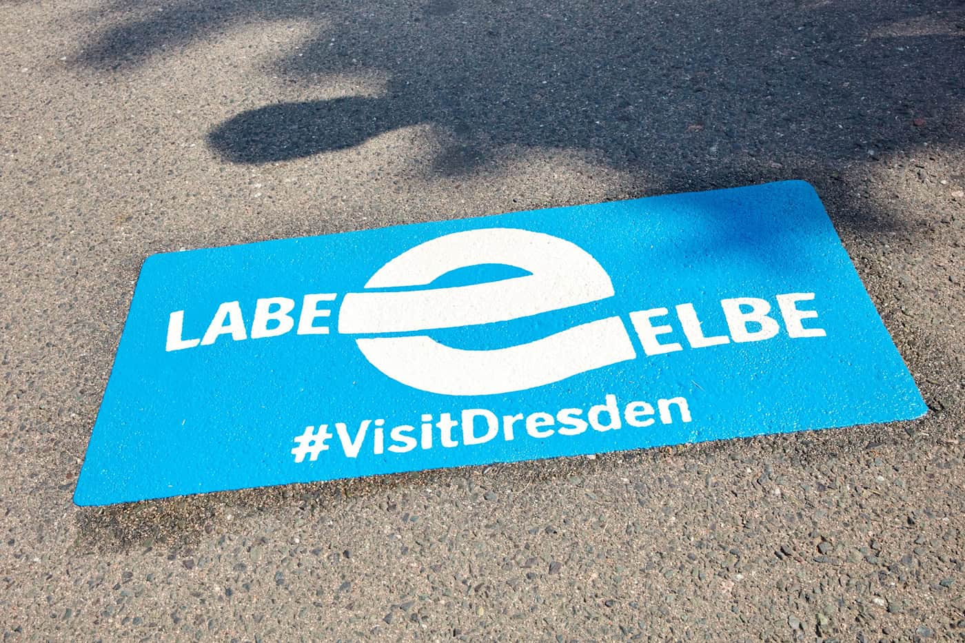 Nahaufnahme eines auf den Asphalt gemalten blauen Rechtecks mit dem weißen Schriftzug Labe Elbe, einem geschwungenen E-Logo und dem Hashtag VisitDresden.