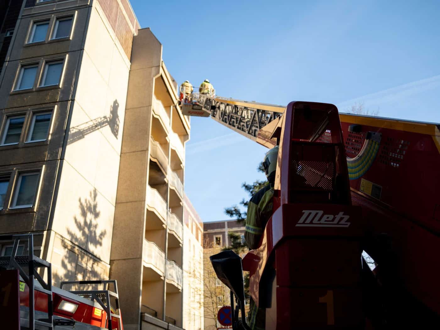 Eine ausgefahrene Drehleiter eines roten Metz-Feuerwehrautos reicht hoch hinauf an der Fassade eines hellen Hochhauses unter blauem Himmel. Zwei Feuerwehrleute stehen im Rettungskorb am oberen Ende. Die Sonne wirft den Schatten der Leiter auf das Gebäude.