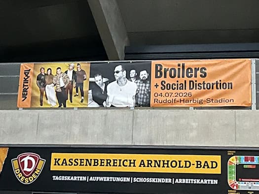 Ein langes, orange-schwarzes Werbebanner für das Broilers-Konzert hängt unter der Betonkonstruktion des Stadions. Darunter befindet sich ein gelb-schwarzes Schild mit der Aufschrift „Kassenbereich Arnhold-Bad“ und dem Logo von Dynamo Dresden.