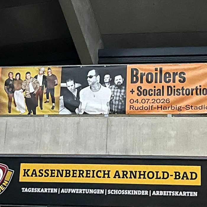 Ein langes, orange-schwarzes Werbebanner für das Broilers-Konzert hängt unter der Betonkonstruktion des Stadions. Darunter befindet sich ein gelb-schwarzes Schild mit der Aufschrift „Kassenbereich Arnhold-Bad“ und dem Logo von Dynamo Dresden.