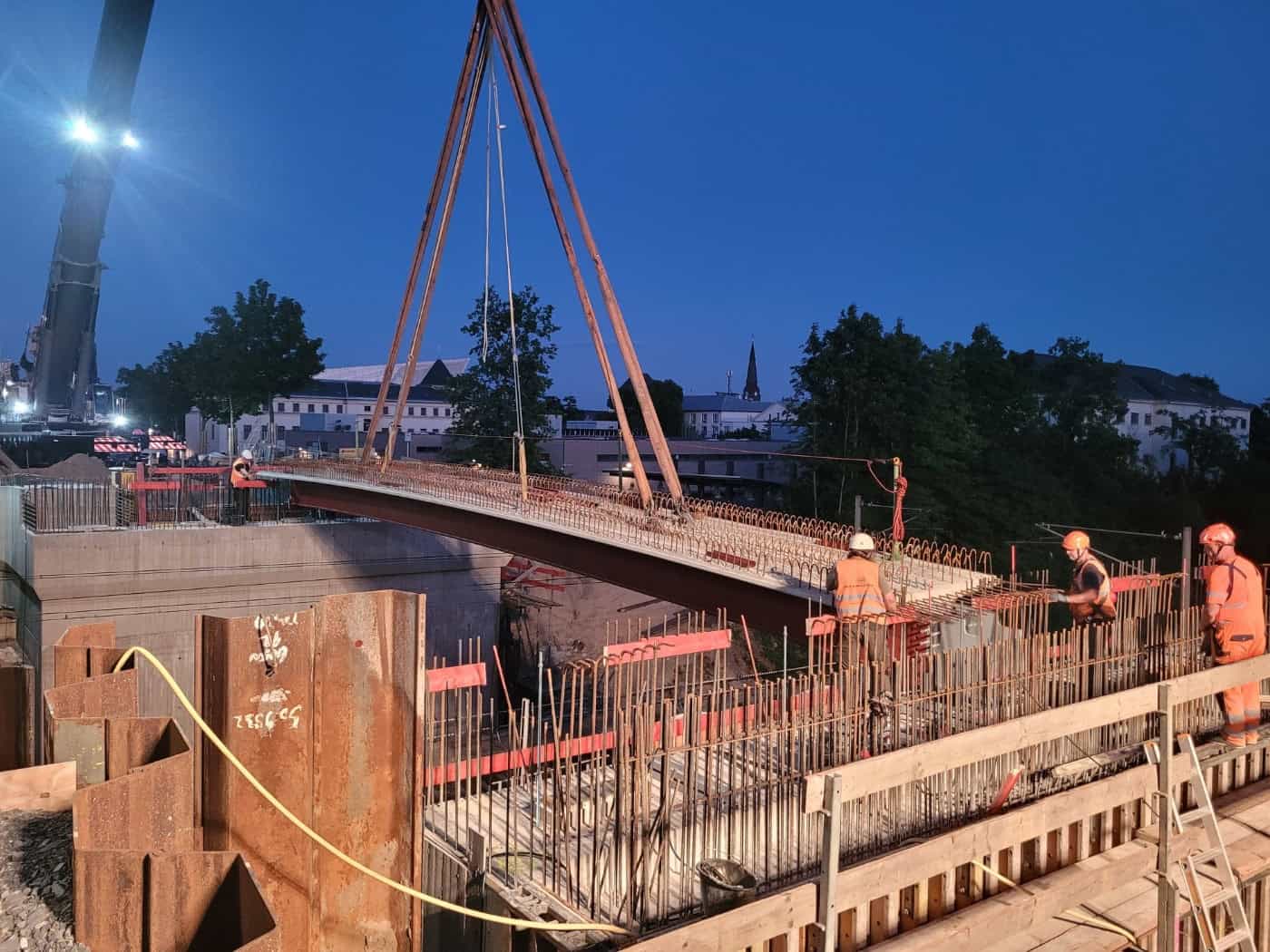 Bauarbeiter in orangefarbener Warnkleidung sichern auf einer Baustelle bei Dämmerung einen massiven Stahlträger, der an Seilen eines großen Krans über einer Betonkonstruktion mit hervorstehendem Bewehrungsstahl schwebt.