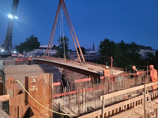 Bauarbeiter in orangefarbener Warnkleidung sichern auf einer Baustelle bei Dämmerung einen massiven Stahlträger, der an Seilen eines großen Krans über einer Betonkonstruktion mit hervorstehendem Bewehrungsstahl schwebt.