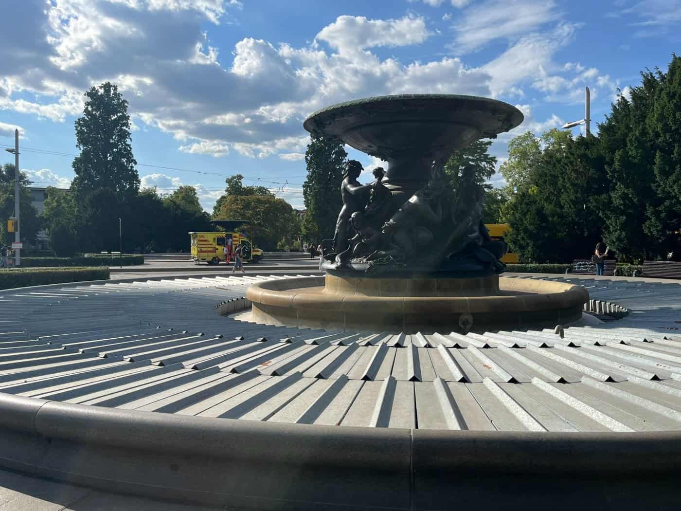Ein historischer Brunnen mit dunklen Figurenskulpturen ist mit einer grauen Metallabdeckung verschlossen. Dahinter erstreckt sich ein Platz mit grünen Bäumen, auf dem ein gelb-rot gestreifter Rettungswagen der Feuerwehr mit Blaulichtaufbau steht.