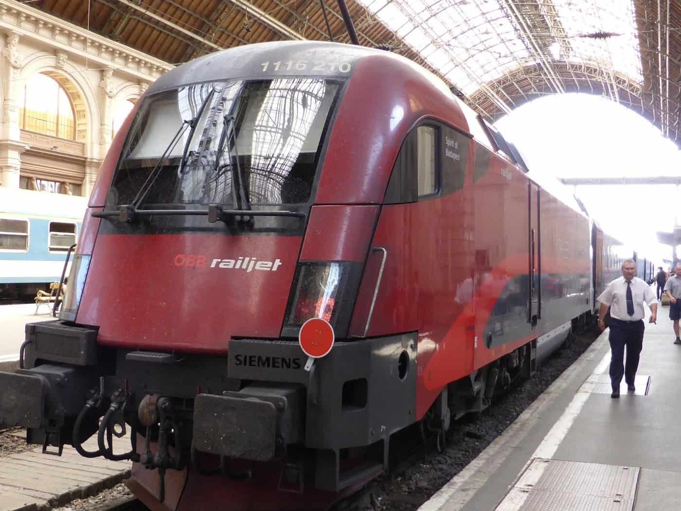 Nahaufnahme der markanten, abgerundeten roten Front eines ÖBB Railjet Zuges in einer historischen Bahnhofshalle mit gewölbtem Glasdach. Rechts auf dem grauen Bahnsteig läuft ein Bahnmitarbeiter in weißem Hemd und dunkler Krawatte am Zug vorbei.