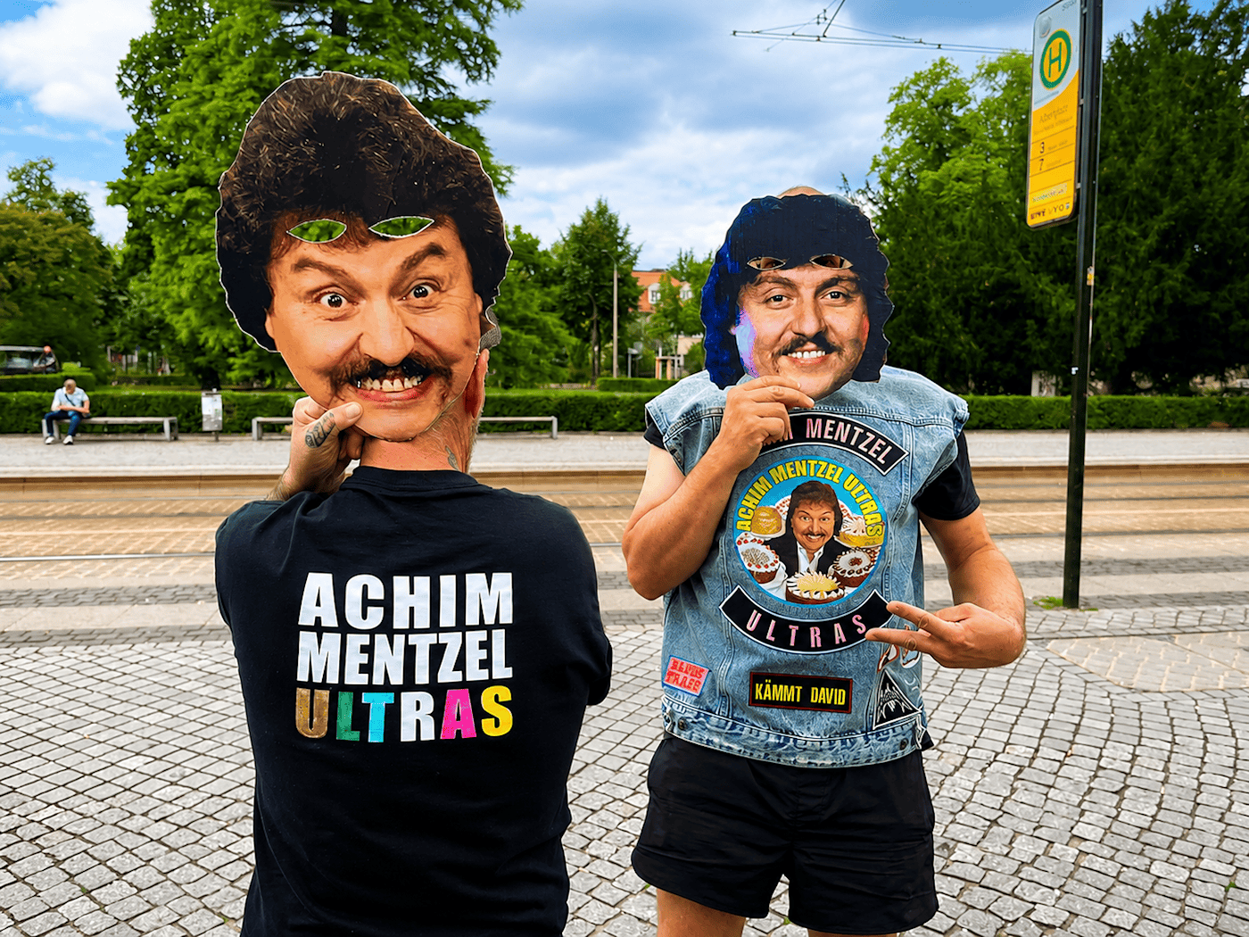 Zwei Mitglieder der Achim-Mentzel-Ultras mit Masken und Fan-Kleidung am Albertplatz in Dresden Zwei Männer auf Pflastersteinen halten Pappmasken von Achim Mentzel vor ihre Gesichter. Der linke trägt ein „Achim Mentzel Ultras“-Shirt von hinten, der rechte eine Jeansweste mit Fan-Aufnähern. Im Hintergrund grüne Bäume und ein gelbes Haltestellenschild.
