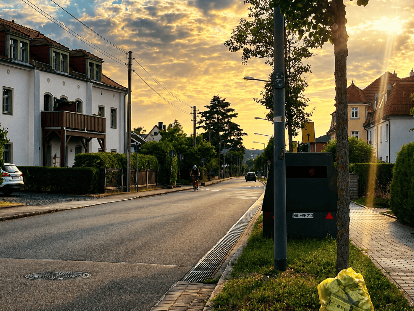 Ein dunkelgrüner, kastenförmiger Blitzer-Anhänger mit dem Kennzeichen HWI-VE 213 steht am rechten Rand einer Wohnstraße hinter einem Baum. Links sind helle Wohnhäuser zu sehen, im Vordergrund auf dem Grünstreifen liegt ein prall gefüllter gelber Sack.