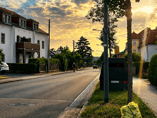 Ein dunkelgrüner, kastenförmiger Blitzer-Anhänger mit dem Kennzeichen HWI-VE 213 steht am rechten Rand einer Wohnstraße hinter einem Baum. Links sind helle Wohnhäuser zu sehen, im Vordergrund auf dem Grünstreifen liegt ein prall gefüllter gelber Sack.