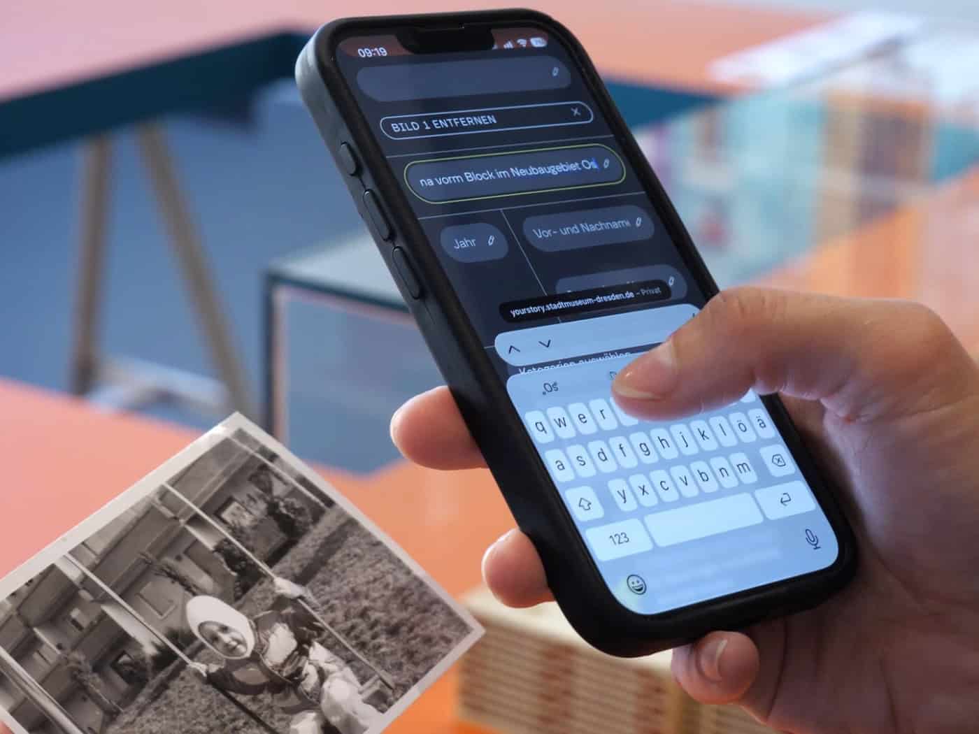 Eine Hand hält ein Smartphone, auf dem Text in ein Online-Formular eingetippt wird. Die andere Hand hält ein altes Schwarz-Weiß-Foto eines Kindes vor einem Plattenbaugebäude.