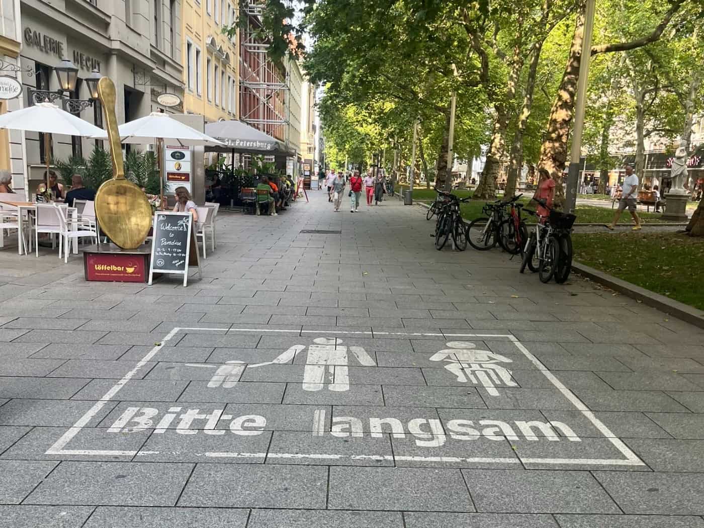 Ein deutliches weißes Piktogramm auf grauem Pflaster zeigt Fußgänger, ein Fahrrad und den Schriftzug „Bitte langsam“. Links befindet sich die Außengastronomie eines Cafés mit einem großen goldenen Löffel als Deko.