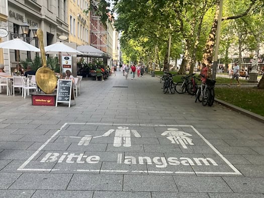 Ein deutliches weißes Piktogramm auf grauem Pflaster zeigt Fußgänger, ein Fahrrad und den Schriftzug „Bitte langsam“. Links befindet sich die Außengastronomie eines Cafés mit einem großen goldenen Löffel als Deko.
