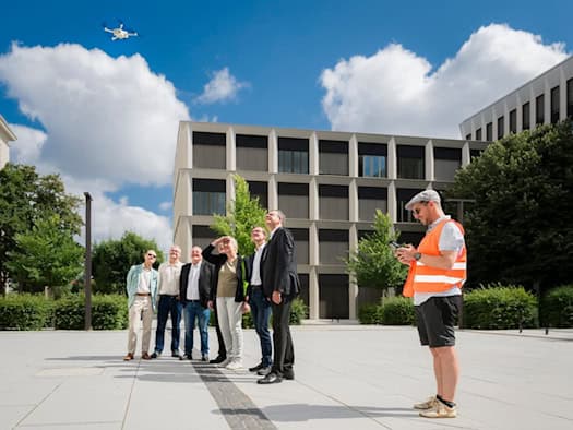 Ein Mann in oranger Warnweste bedient auf einem hellen Platz eine Drohne, die am Himmel fliegt. Links steht eine Gruppe von sieben Personen und blickt nach oben. Im Hintergrund befindet sich ein modernes, rasterförmiges Gebäude.