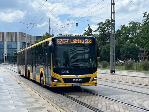 Ein gelb-schwarzer MAN-Gelenkbus der DVB mit der leuchtenden Digitalanzeige „62 Löbtau Süd“ fährt auf einer gepflasterten Straße mit eingelassenen Straßenbahnschienen. Im Hintergrund stehen moderne und historische Gebäude unter einem leicht bewölkten Himmel.