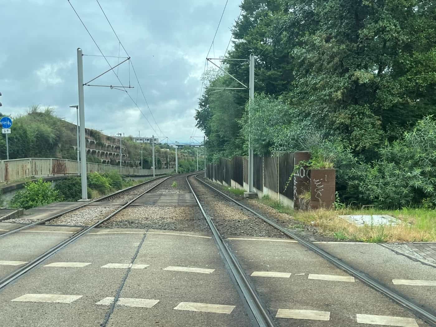 Eine zweigleisige Straßenbahntrasse verläuft in einer Kurve nach rechts, flankiert von einer Betonwand auf der linken Seite und grünen Bäumen sowie einem Holzzaun auf der rechten Seite unter bewölktem Himmel.