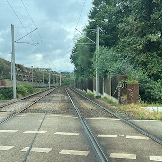Eine zweigleisige Straßenbahntrasse verläuft in einer Kurve nach rechts, flankiert von einer Betonwand auf der linken Seite und grünen Bäumen sowie einem Holzzaun auf der rechten Seite unter bewölktem Himmel.