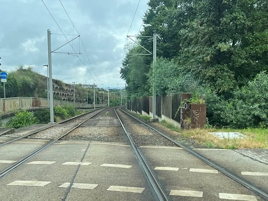 Eine zweigleisige Straßenbahntrasse verläuft in einer Kurve nach rechts, flankiert von einer Betonwand auf der linken Seite und grünen Bäumen sowie einem Holzzaun auf der rechten Seite unter bewölktem Himmel.