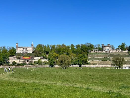 Weite, grüne Elbwiesen im Vordergrund, auf denen links Spaziergänger laufen. Dahinter erhebt sich ein dicht bewaldeter Hang mit zwei prachtvollen historischen Schlössern – Schloss Albrechtsberg links und das Lingnerschloss rechts – unter strahlend blauem Himmel.