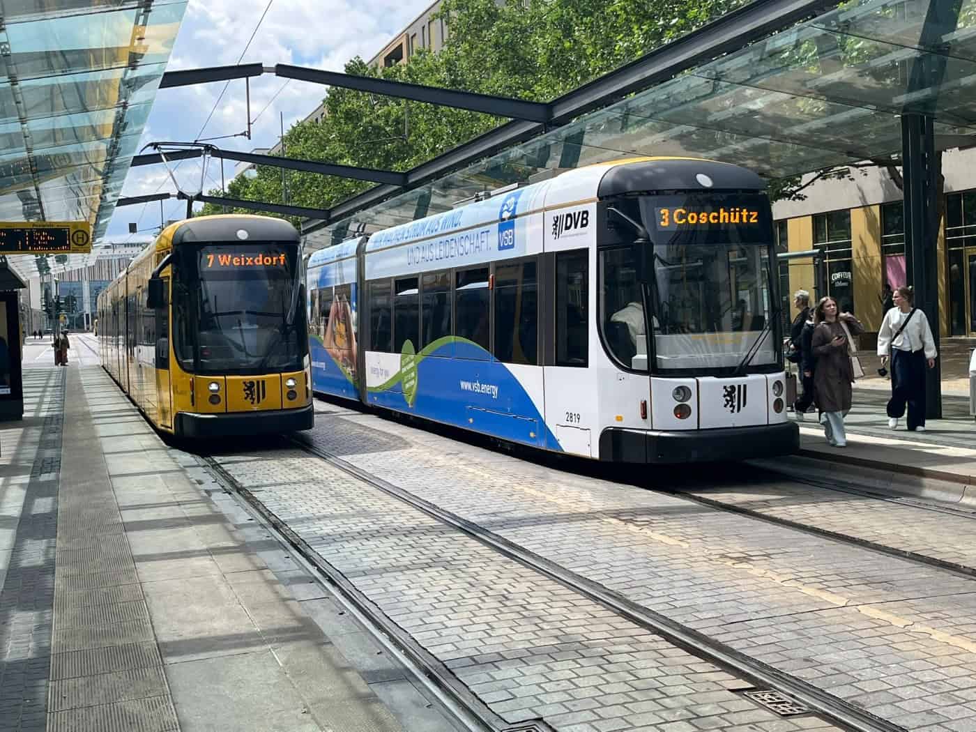 Zwei moderne Straßenbahnen der DVB stehen nebeneinander an einer überdachten Haltestelle. Auf den gelben LED-Anzeigen stehen "7 Weixdorf" und "3 Coschütz". Rechts gehen Passanten auf dem gepflasterten Bahnsteig im Sonnenschein entlang.