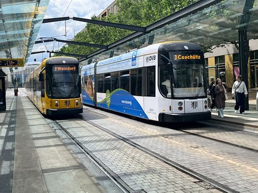 Zwei moderne Straßenbahnen der DVB stehen nebeneinander an einer überdachten Haltestelle. Auf den gelben LED-Anzeigen stehen "7 Weixdorf" und "3 Coschütz". Rechts gehen Passanten auf dem gepflasterten Bahnsteig im Sonnenschein entlang.