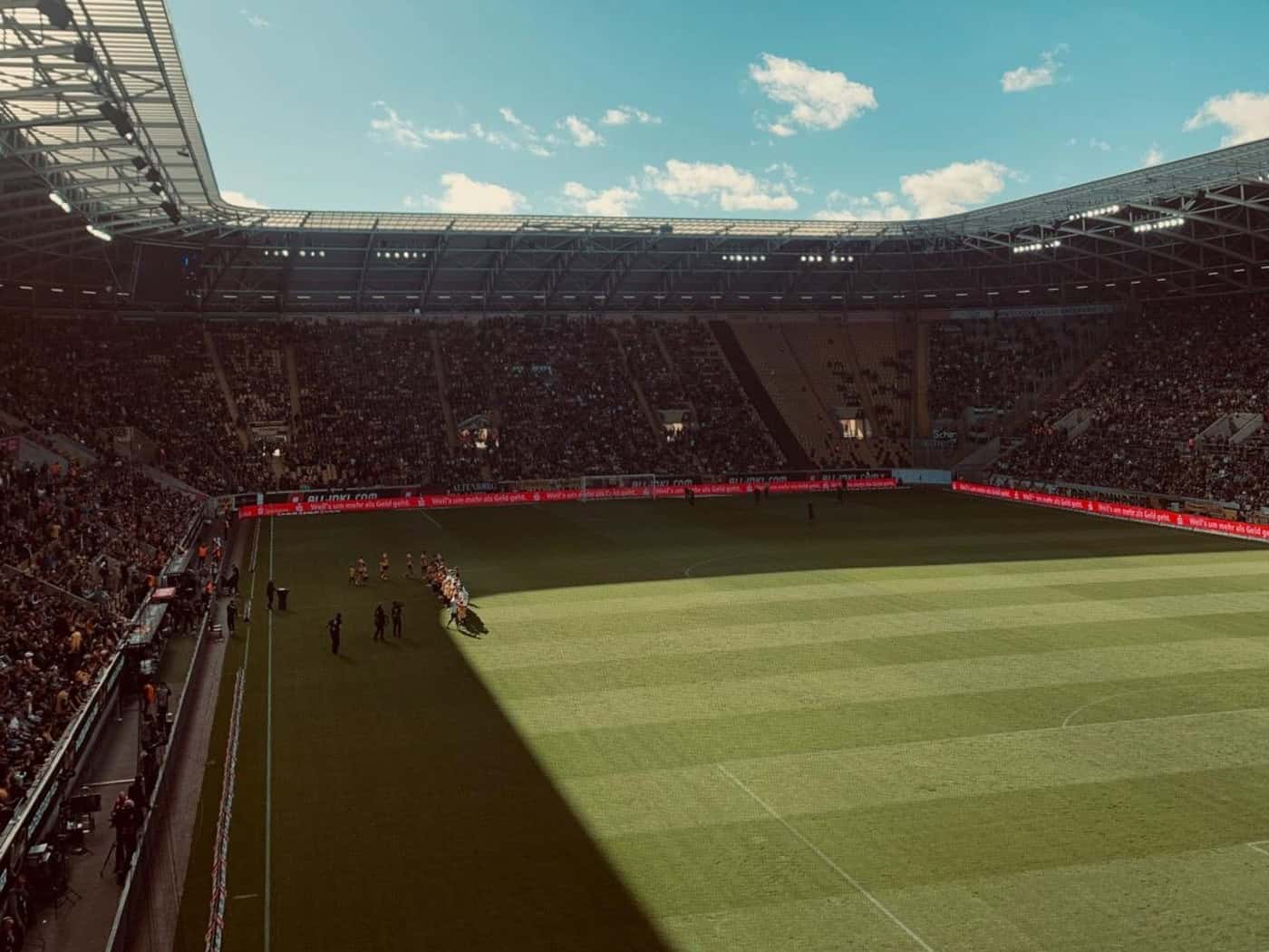 Blick aus erhöhter Perspektive in ein vollbesetztes Fußballstadion mit besetzten Tribünen und grünem Spielfeld. Ein großer Schatten fällt diagonal über den Rasen. Über der offenen Dachkonstruktion ist ein strahlend blauer Himmel mit wenigen Wolken zu sehen.