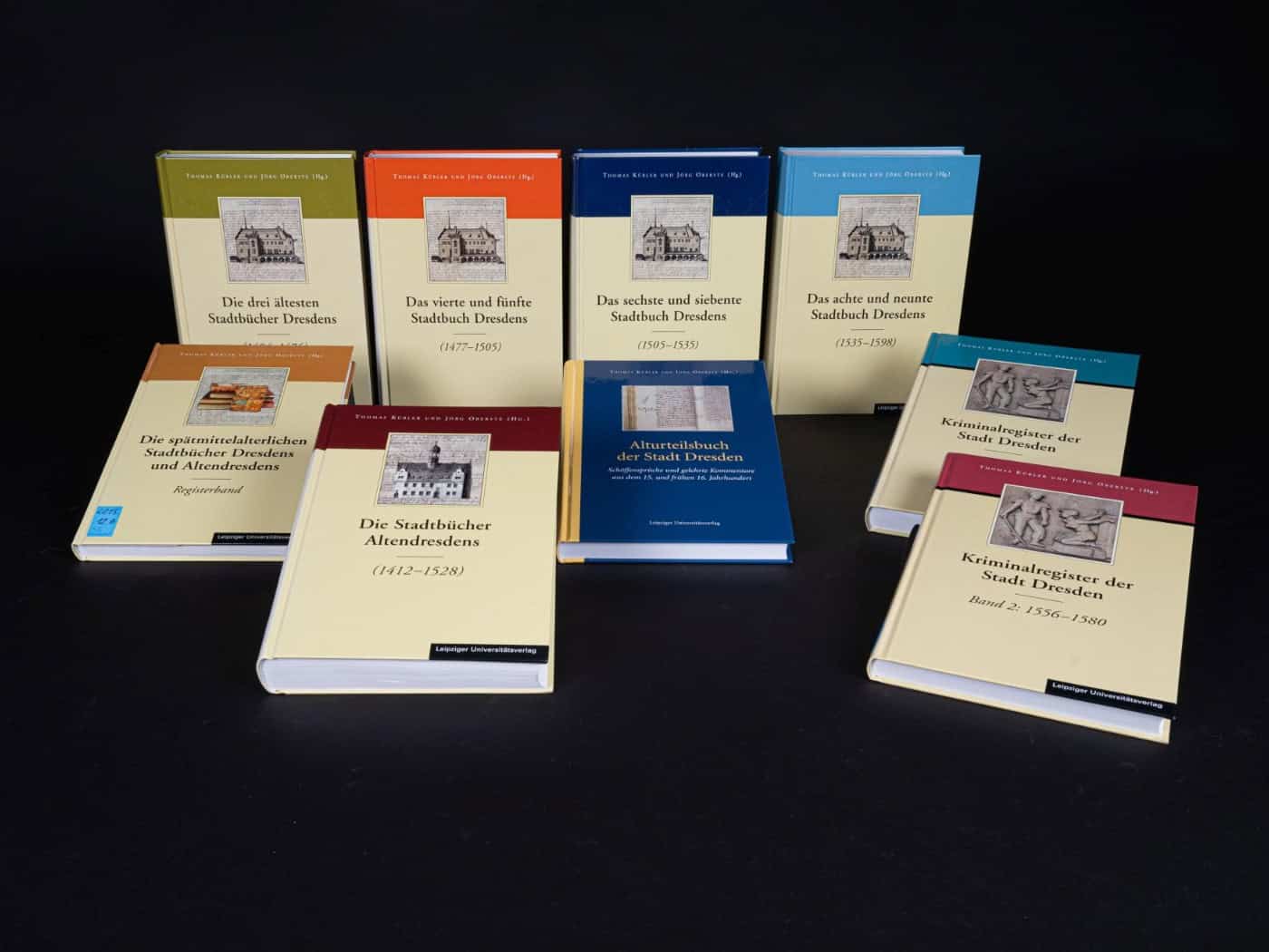 Acht dicke, beige Hardcover-Bücher mit farbigen Kopfleisten in Grün, Orange, Blau und Rot sind in zwei Reihen auf schwarzem Hintergrund aufgestellt. Auf den Einbänden sind historische Grafiken und Titel wie „Kriminalregister der Stadt Dresden“ zu sehen.